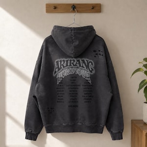 May include: A charcoal gray hooded sweatshirt with a distressed, washed-out finish. The back features the text ARIRANG WORLD TOUR in a stylized font, followed by a list of global city names and the phrase AND MORE. The fabric has small holes.