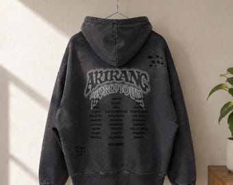 May include: A charcoal gray hooded sweatshirt with a distressed, washed-out finish. The back features the text ARIRANG WORLD TOUR in a bold, arched font, followed by a list of global city names and the phrase AND MORE in black lettering.