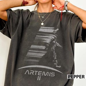 Puede incluir: Camiseta gris oscuro con un gráfico de un astronauta y el texto "ARTEMIS II". El diseño incluye un efecto de rayas diagonales y una línea curva en la parte inferior. La palabra "PEPPER" está en la esquina inferior derecha.