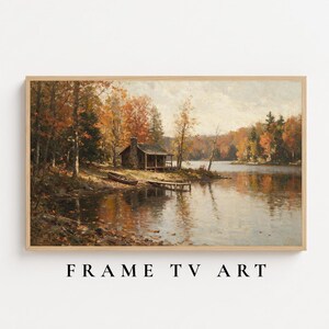 May include: A landscape painting in a light wood frame depicting a rustic cabin on a lake shore surrounded by autumn trees with orange and yellow foliage. A small wooden boat rests on the bank near a dock, with the scene reflected in the calm water.