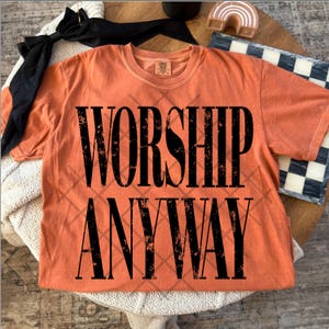 May include: An orange t-shirt with the words "WORSHIP ANYWAY" in large, distressed black letters. The shirt has a crew neck and short sleeves. A black ribbon and decorative items are in the background.