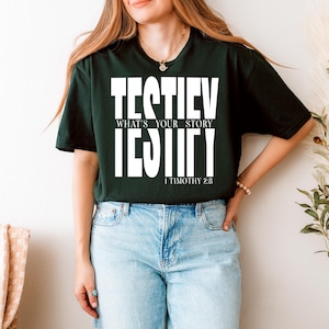 Pode incluir: T-shirt verde escuro com o texto branco "TESTIFY WHAT'S YOUR STORY IDENTIFY" e "1 TIMOTHY 2:8". A t-shirt é combinada com jeans azul claro. O design da t-shirt é uma declaração gráfica arrojada.
