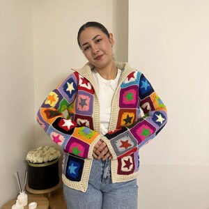 May include: A colorful, crocheted cardigan with a star pattern. The jacket features a cream-colored collar and trim, and a zipper closure. The squares are in various colors, including blue, green, pink, and yellow. The cardigan is a unique, handmade piece.