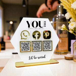 Könnte beinhalten: Ein weißes und goldenes Acrylschild mit dem Wort "YOU" oben, gefolgt von "beauty | ink | health". Es zeigt goldene Symbole für WhatsApp, Facebook und Pinterest, jeweils mit einem entsprechenden QR-Code. Der Satz "Let be social" steht unten.