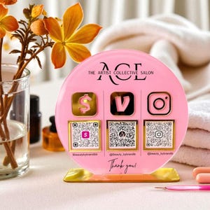 May include: A pink acrylic salon sign with the text "ACE THE ARTIST COLLECTIVE SALON". The sign has three social media icons with QR codes and the words "Thank you!". The base is gold.
