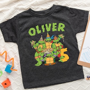 May include: Dark gray t-shirt featuring a graphic of the Teenage Mutant Ninja Turtles holding pizza, with the name "OLIVER" in green above and the number "5" below. The shirt is on a white textured surface.