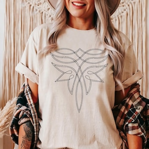 May include: A beige t-shirt with a black embroidered design of a stylized geometric pattern. The design resembles a stylized flower or a geometric mandala.