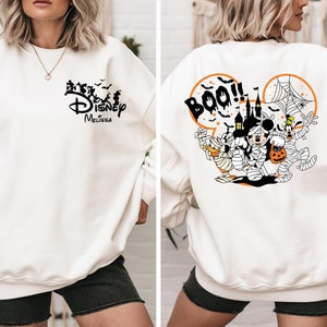 May include: Off-white sweatshirt with a Halloween-themed Disney graphic. The front features the Disney logo and silhouettes. The back displays Mickey Mouse, Donald Duck, and Goofy as mummies, with a haunted house and the word "Boo!!" in an orange circle.