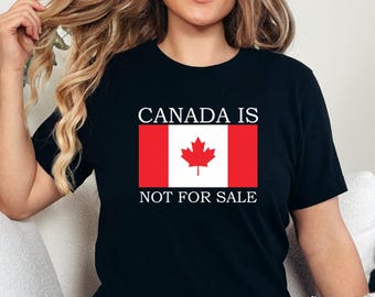May include: Black t-shirt featuring the Canadian flag with the text "CANADA IS NOT FOR SALE" in white. The flag design includes a red maple leaf on a white background, framed by red vertical bars. The shirt is a casual wear item.