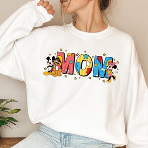 Peut inclure: Sweat-shirt blanc avec le mot "MOM" en lettres color&eacute;es et des &eacute;toiles. Mickey Mouse, Pluto et Minnie Mouse sont repr&eacute;sent&eacute;s sur le sweat-shirt, ajoutant une touche ludique au design.