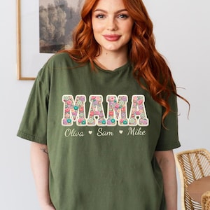 May include: An olive green t-shirt with the word "MAMA" in a floral design. Below the word "MAMA" are the names "Olivia", "Sam", and "Mike". The shirt is a short-sleeved, crew neck style.
