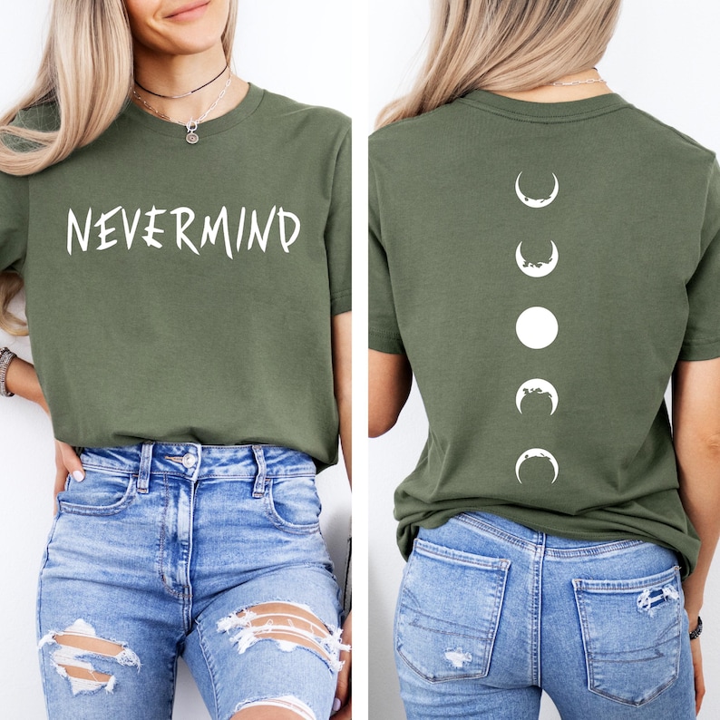 May include: Olive green t-shirt with the word "NEVERMIND" in white on the front. The back of the shirt features a vertical design of white moon phases. The shirt is paired with blue jeans.