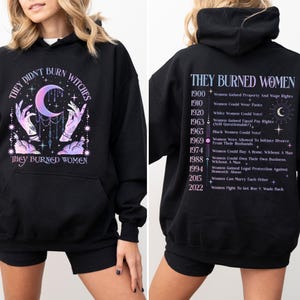May include: Black hooded sweatshirt with a graphic print of hands holding a crescent moon and stars. The back of the sweatshirt has text that reads "They Burned Women" and lists historical events related to women's rights in the United States.