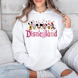 May include: White hooded sweatshirt with a pink Disneyland graphic featuring Mickey Mouse, Minnie Mouse, Donald Duck, Goofy, and Pluto. The graphic also includes pink hearts and fireworks.