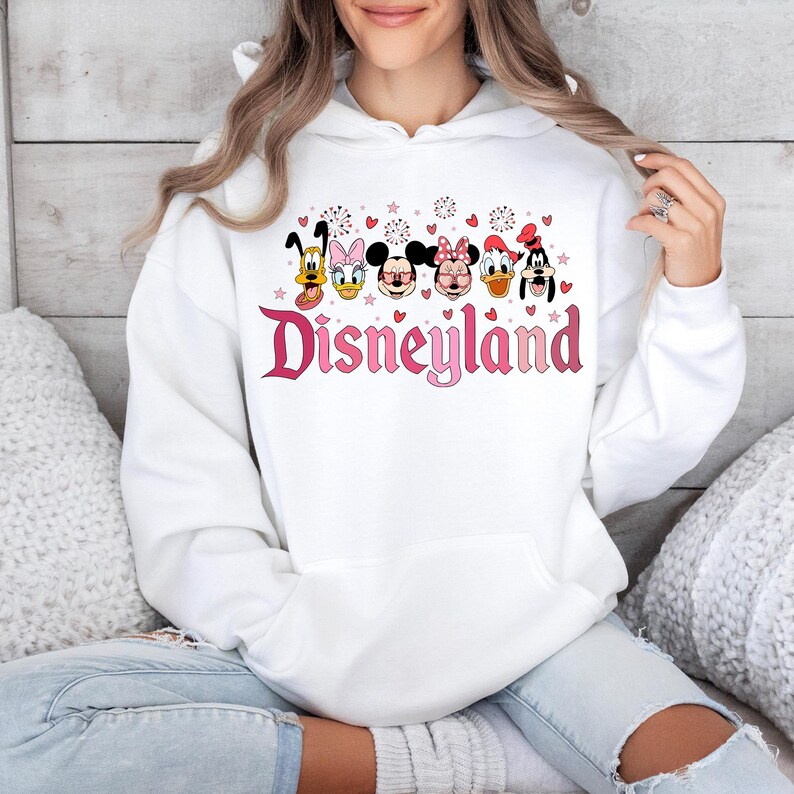 May include: White hooded sweatshirt with a pink Disneyland graphic featuring Mickey Mouse, Minnie Mouse, Donald Duck, Goofy, and Pluto. The graphic also includes pink hearts and fireworks.