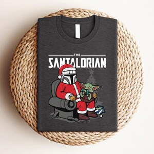 May include: Dark gray t-shirt featuring a festive design. The graphic shows a Mandalorian character in a Santa Claus outfit, seated with a small green alien. The text reads "THE SANTALORIAN" in white letters.