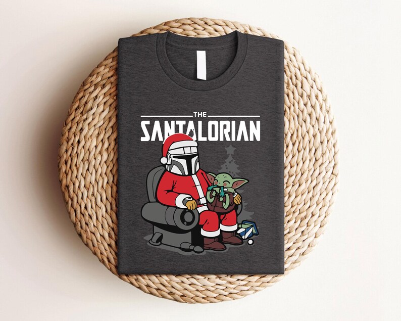 May include: Dark gray t-shirt featuring a festive design. The graphic shows a Mandalorian character in a Santa Claus outfit, seated with a small green alien. The text reads "THE SANTALORIAN" in white letters.