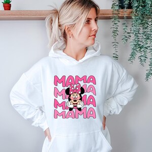 K&ouml;nnte beinhalten: Wei&szlig;er Kapuzenpullover mit einem rosa Aufdruck, der das Wort "MAMA" viermal wiederholt, mit einem Minnie-Mouse-Design in der Mitte. Das Minnie-Mouse-Design hat eine rosa Schleife und ein l&auml;chelndes Gesicht. Der Kapuzenpullover hat eine Vordertasche und eine Kapuze mit Kordelzug.