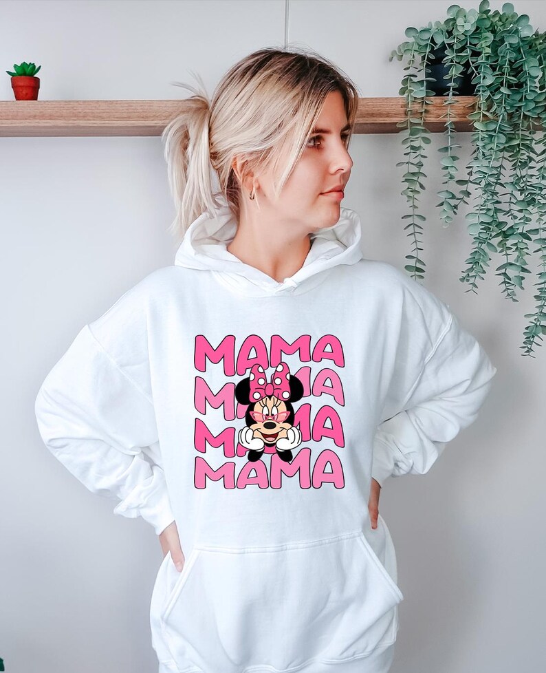 K&ouml;nnte beinhalten: Wei&szlig;er Kapuzenpullover mit einem rosa Aufdruck, der das Wort "MAMA" viermal wiederholt, mit einem Minnie-Mouse-Design in der Mitte. Das Minnie-Mouse-Design hat eine rosa Schleife und ein l&auml;chelndes Gesicht. Der Kapuzenpullover hat eine Vordertasche und eine Kapuze mit Kordelzug.