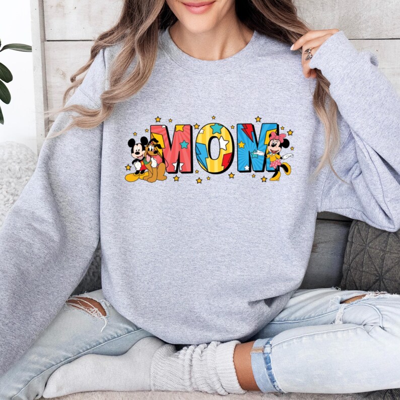 Peut inclure: Un sweat-shirt gris clair avec le mot "MOM" en lettres color&eacute;es de style dessin anim&eacute;. Mickey Mouse, Pluto et Minnie Mouse sont repr&eacute;sent&eacute;s sur le sweat-shirt. Le sweat-shirt est de style col rond.