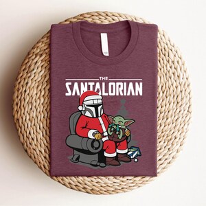 May include: Burgundy t-shirt featuring a festive graphic. The design includes the text "THE SANTALORIAN" above an illustration of a character in a Santa suit with a helmet, seated with a green alien. A fun holiday themed tee.