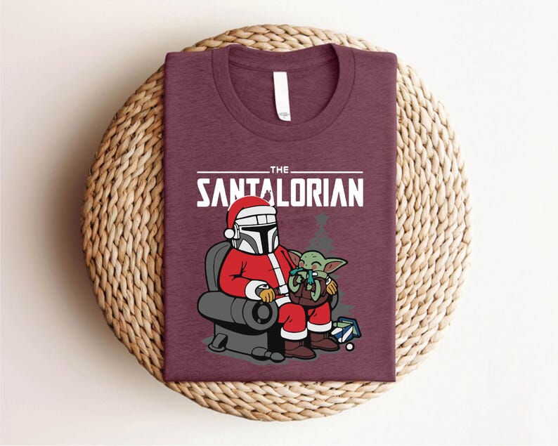 May include: Burgundy t-shirt featuring a festive graphic. The design includes the text "THE SANTALORIAN" above an illustration of a character in a Santa suit with a helmet, seated with a green alien. A fun holiday themed tee.