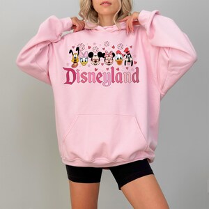 May include: Pink hooded sweatshirt with a graphic print of Mickey Mouse, Minnie Mouse, Donald Duck, Goofy, and Pluto. The graphic also includes the text "Disneyland" in pink and red.