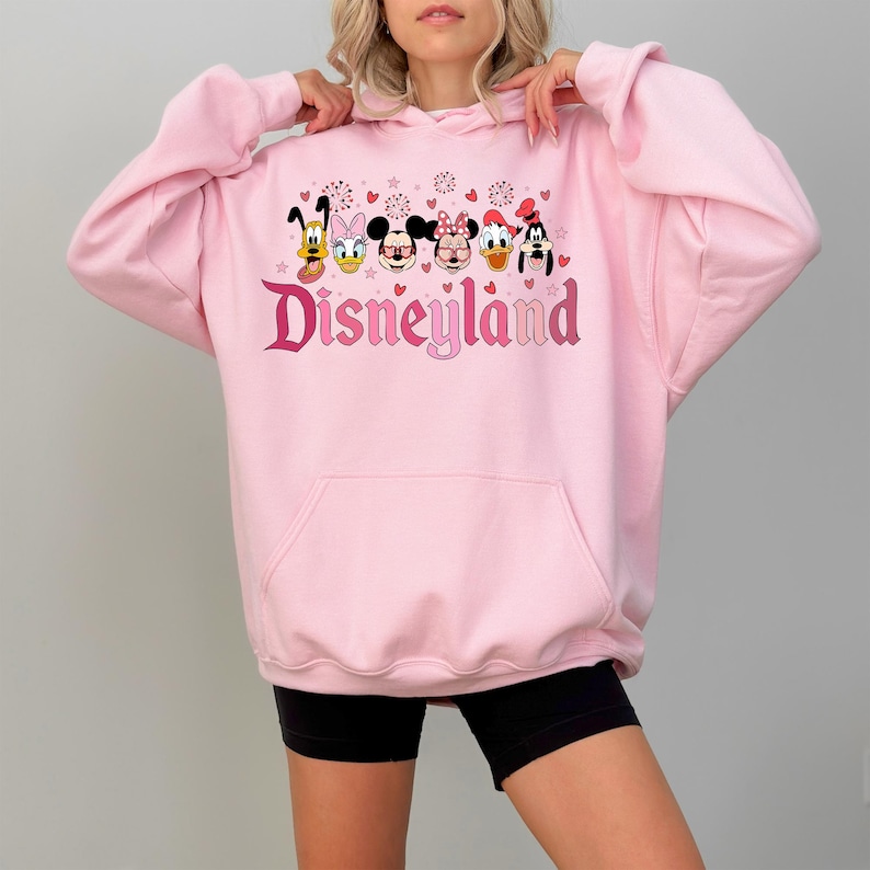 May include: Pink hooded sweatshirt with a graphic print of Mickey Mouse, Minnie Mouse, Donald Duck, Goofy, and Pluto. The graphic also includes the text "Disneyland" in pink and red.