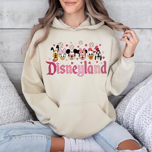 May include: A cream-colored hoodie with a pink Disneyland logo and a row of cartoon characters including Mickey Mouse, Minnie Mouse, Donald Duck, Goofy, and Pluto. The characters are wearing pink hearts and there are pink fireworks above them.