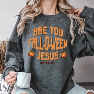 May include: Dark gray sweatshirt with the text "ARE YOU FALL-O-WEEN JESUS" in orange, along with two orange pumpkins. The text "MATTHEW 4:19" is below the main text. A seasonal, fall-themed design.