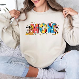 Peut inclure: Sweat-shirt cr&egrave;me avec le mot "MOM" en lettres color&eacute;es de style dessin anim&eacute;. Mickey Mouse, Pluto et Minnie Mouse sont repr&eacute;sent&eacute;s dans le design. De petites &eacute;toiles jaunes entourent le mot "MOM".