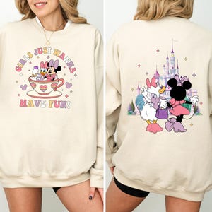 May include: Two cream-colored sweatshirts with different Disney character designs. The first sweatshirt features Mickey Mouse and Daisy Duck in a teacup with the text "Girls Just Wanna Have Fun!" The second sweatshirt features Daisy Duck and Minnie Mouse in front of a pink castle with stars.