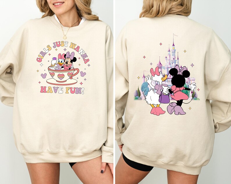 May include: Two cream-colored sweatshirts with different Disney character designs. The first sweatshirt features Mickey Mouse and Daisy Duck in a teacup with the text "Girls Just Wanna Have Fun!" The second sweatshirt features Daisy Duck and Minnie Mouse in front of a pink castle with stars.