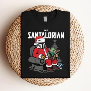 May include: Black t-shirt featuring a cartoon illustration of a character in a Santa Claus outfit, seated with a green alien. The text "THE SANTALORIAN" is printed across the top in white. A Christmas tree and gifts are in the background.