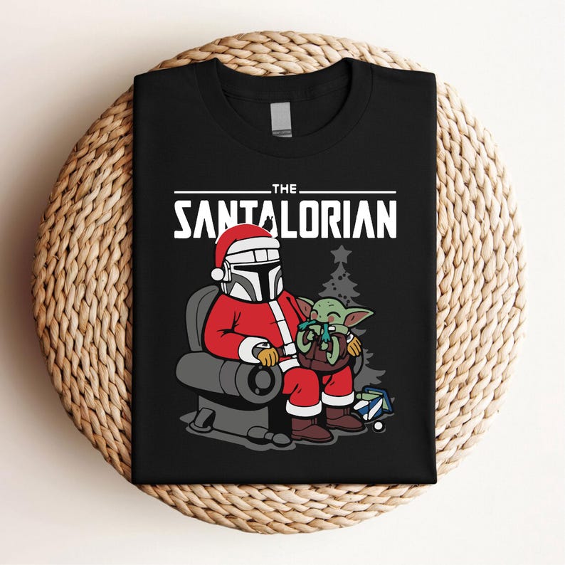 May include: Black t-shirt featuring a cartoon illustration of a character in a Santa Claus outfit, seated with a green alien. The text "THE SANTALORIAN" is printed across the top in white. A Christmas tree and gifts are in the background.