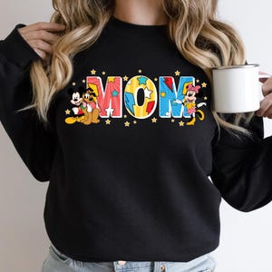 Peut inclure: Sweat-shirt noir avec le mot "MOM" en lettres color&eacute;es et &eacute;toil&eacute;es. Mickey Mouse, Pluto et Minnie Mouse sont repr&eacute;sent&eacute;s sur le sweat-shirt. Une tasse blanche est tenue dans la main de la personne.