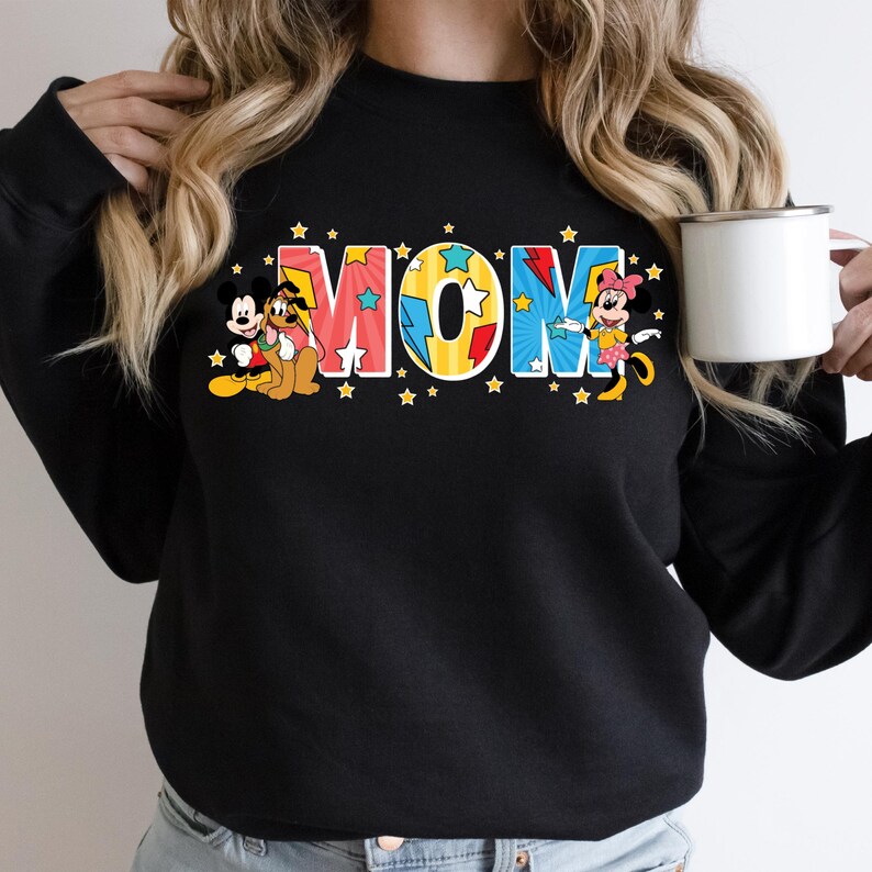 Peut inclure: Sweat-shirt noir avec le mot "MOM" en lettres color&eacute;es et &eacute;toil&eacute;es. Mickey Mouse, Pluto et Minnie Mouse sont repr&eacute;sent&eacute;s sur le sweat-shirt. Une tasse blanche est tenue dans la main de la personne.