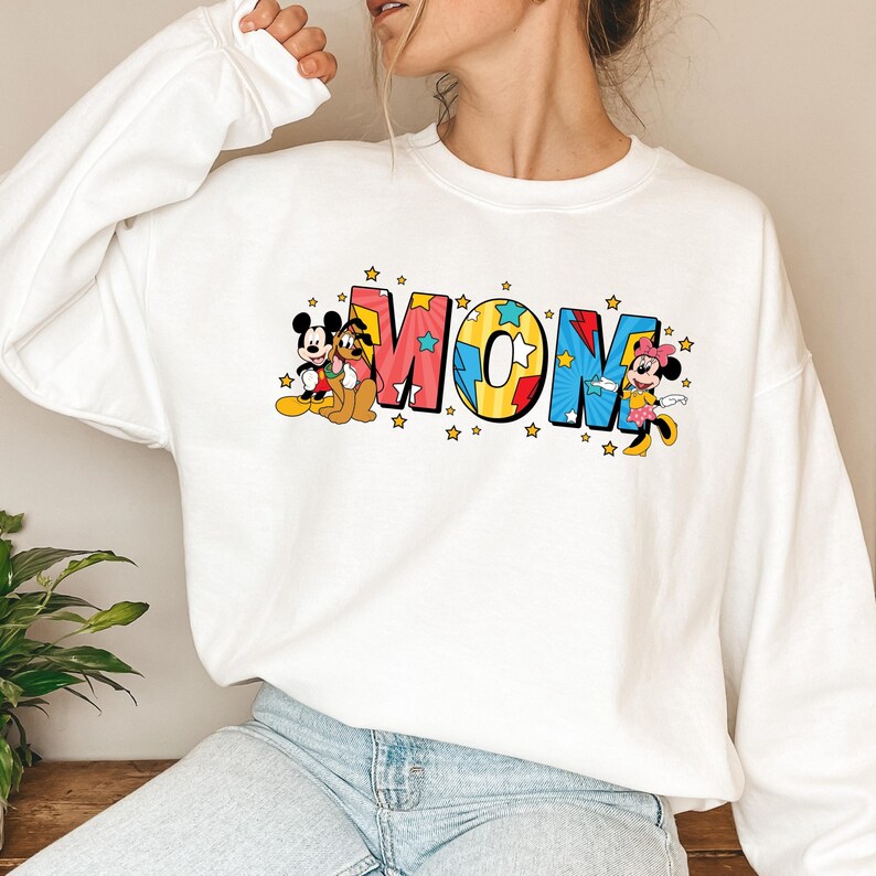 Peut inclure: Sweat-shirt blanc avec le mot "MOM" en lettres color&eacute;es et des &eacute;toiles. Mickey Mouse, Pluto et Minnie Mouse sont repr&eacute;sent&eacute;s sur le sweat-shirt, ajoutant une touche ludique au design.