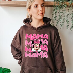 K&ouml;nnte beinhalten: Ein brauner Kapuzenpullover mit einer Vordertasche. Auf der Vorderseite steht das Wort "MAMA" in Rosa, dreifach gestapelt, mit einer Minnie Mouse-Grafik in der Mitte.