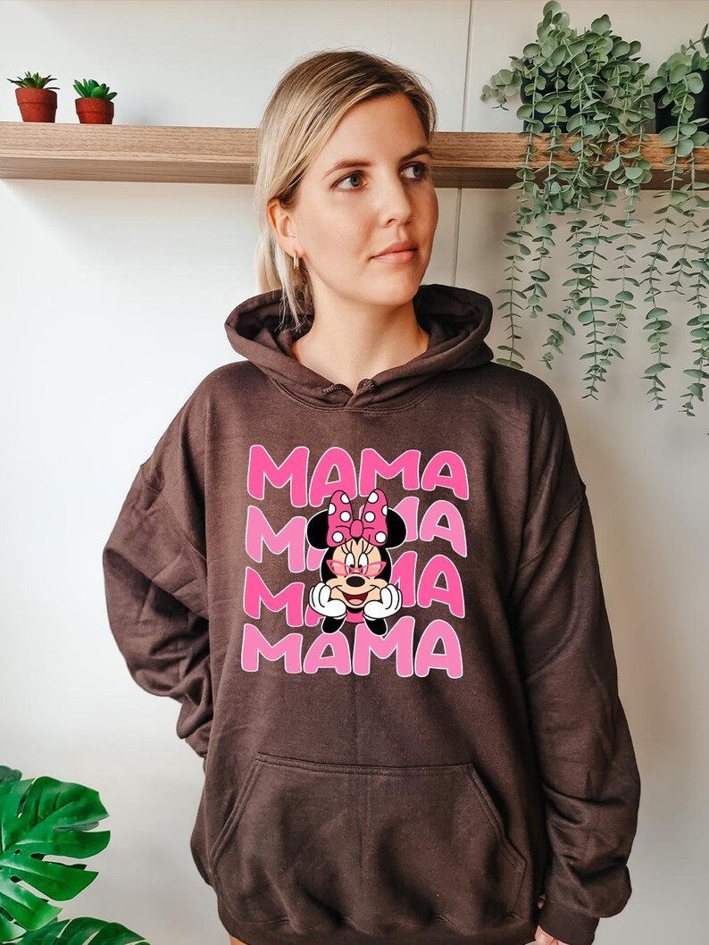 K&ouml;nnte beinhalten: Ein brauner Kapuzenpullover mit einer Vordertasche. Auf der Vorderseite steht das Wort "MAMA" in Rosa, dreifach gestapelt, mit einer Minnie Mouse-Grafik in der Mitte.