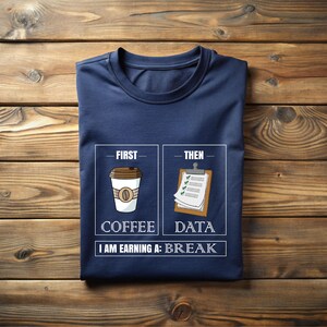 May include: Navy blue t-shirt with a graphic that reads "First Coffee, Then Data, I Am Earning A: Break." The design features a coffee cup and a clipboard. The shirt is folded on a wooden surface.