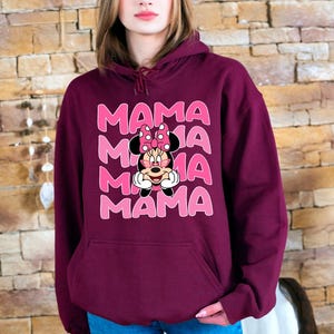 K&ouml;nnte beinhalten: Ein weinroter Kapuzenpullover mit einer Vordertasche und einer Grafik von Minnie Mouse und dem Wort "MAMA" in Rosa. Der Kapuzenpullover ist ein Pullover-Stil und besteht aus einem weichen Material.