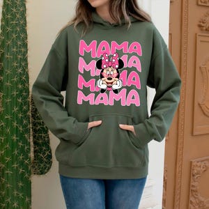 K&ouml;nnte beinhalten: Ein olivgr&uuml;ner Kapuzenpullover mit dem Wort "MAMA" dreimal in Rosa gestapelt, mit einer Minnie Mouse-Grafik in der Mitte. Der Kapuzenpullover hat eine Vordertasche und wird mit blauen Jeans getragen.