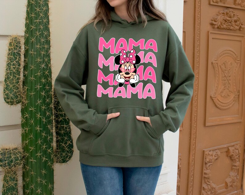 K&ouml;nnte beinhalten: Ein olivgr&uuml;ner Kapuzenpullover mit dem Wort "MAMA" dreimal in Rosa gestapelt, mit einer Minnie Mouse-Grafik in der Mitte. Der Kapuzenpullover hat eine Vordertasche und wird mit blauen Jeans getragen.