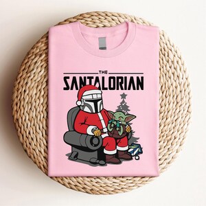 May include: Pink t-shirt with the text "THE SANTALORIAN" in black. The graphic shows a character in a Santa hat and suit, seated, holding a green alien. A Christmas tree and gifts are also in the image.