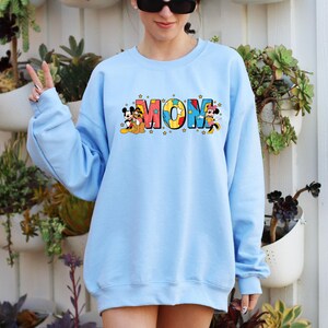 Peut inclure: Un sweat-shirt bleu clair avec le mot "MOM" en lettres color&eacute;es, accompagn&eacute; de graphiques de Mickey et Minnie Mouse. Le sweat-shirt a un col rond et des manches longues. Le design comprend des &eacute;toiles.