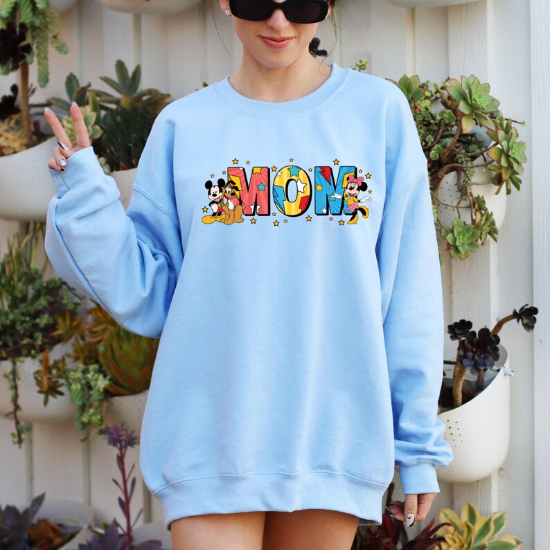 Peut inclure: Un sweat-shirt bleu clair avec le mot "MOM" en lettres color&eacute;es, accompagn&eacute; de graphiques de Mickey et Minnie Mouse. Le sweat-shirt a un col rond et des manches longues. Le design comprend des &eacute;toiles.