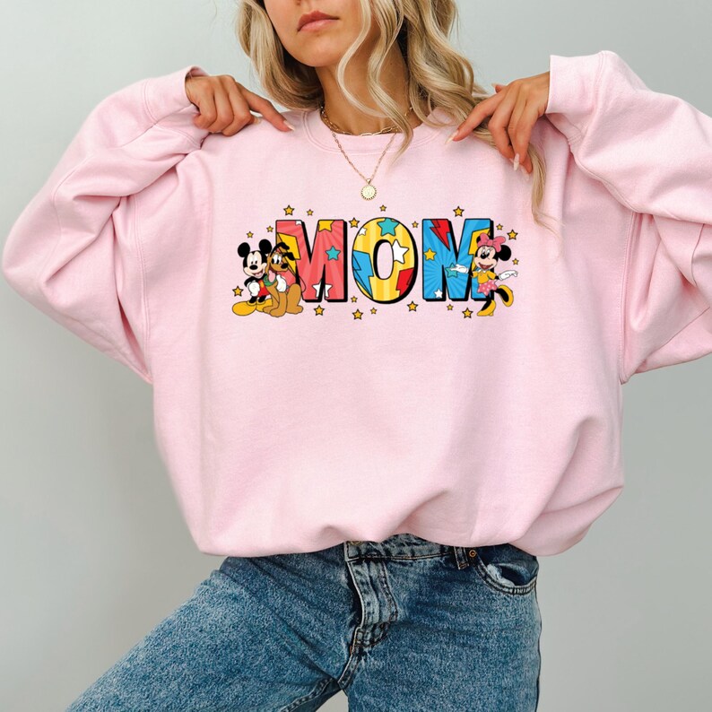 Peut inclure: Un sweat-shirt rose clair avec le mot "MOM" en lettres color&eacute;es, accompagn&eacute; de graphiques de Mickey Mouse, Dingo et Minnie Mouse. Le sweat-shirt est de style col rond et est fait d'une mati&egrave;re douce.