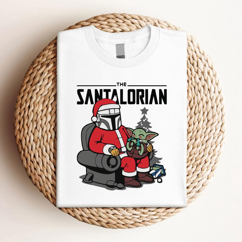 May include: White t-shirt featuring a festive design. The graphic depicts a character in a Santa Claus outfit with a baby alien. The text "THE SANTALORIAN" is printed above the characters. A Christmas tree and a gift box are also included.
