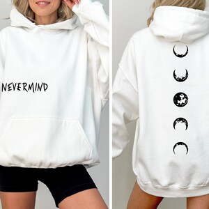 May include: White hoodie with a front pocket and the word "NEVERMIND" printed in black. The back of the hoodie features a series of moon phase graphics in black. A casual, comfortable garment.